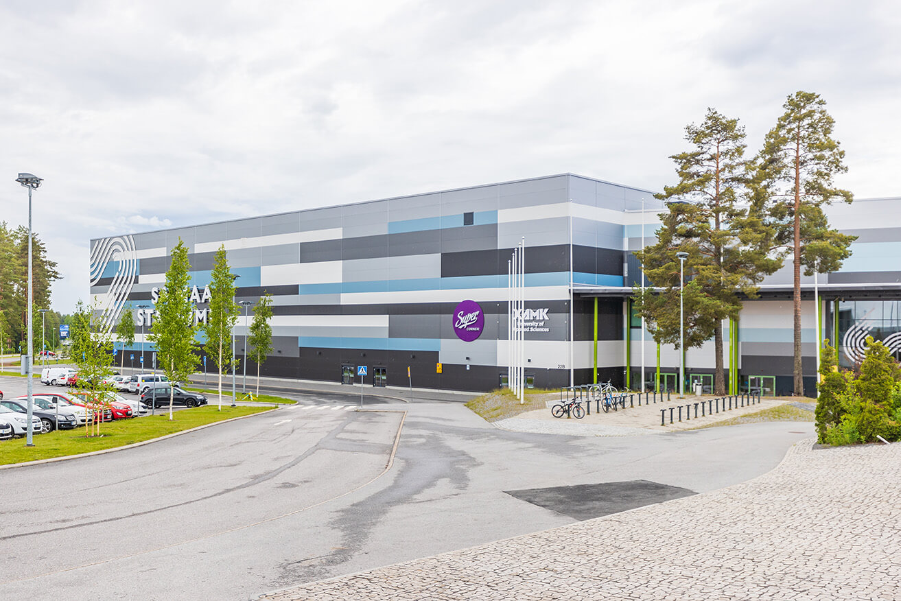 Asfalttitie ja parkkipaikka suuren rakennuksen edustalla. 